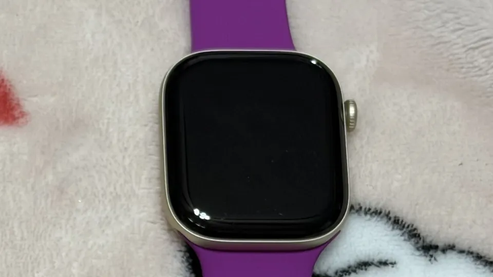 Apple Watch Series 12 in light gold aluminum with a magenta Sport Band, screen off, lying on a soft blanket