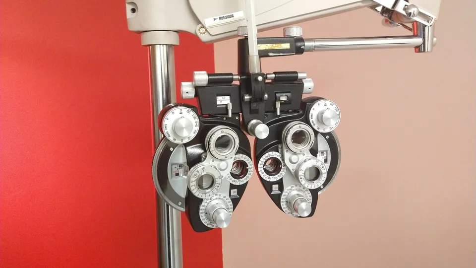 A phoropter, an eye-exam instrument with rows of lens dials, hanging from a mounted arm in front of a red and beige wall