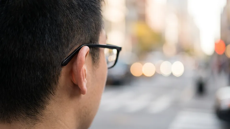 The back of a person's head and ear with the thin temple arm of plain black glasses resting above the ear, a blurred city street with warm lights behind