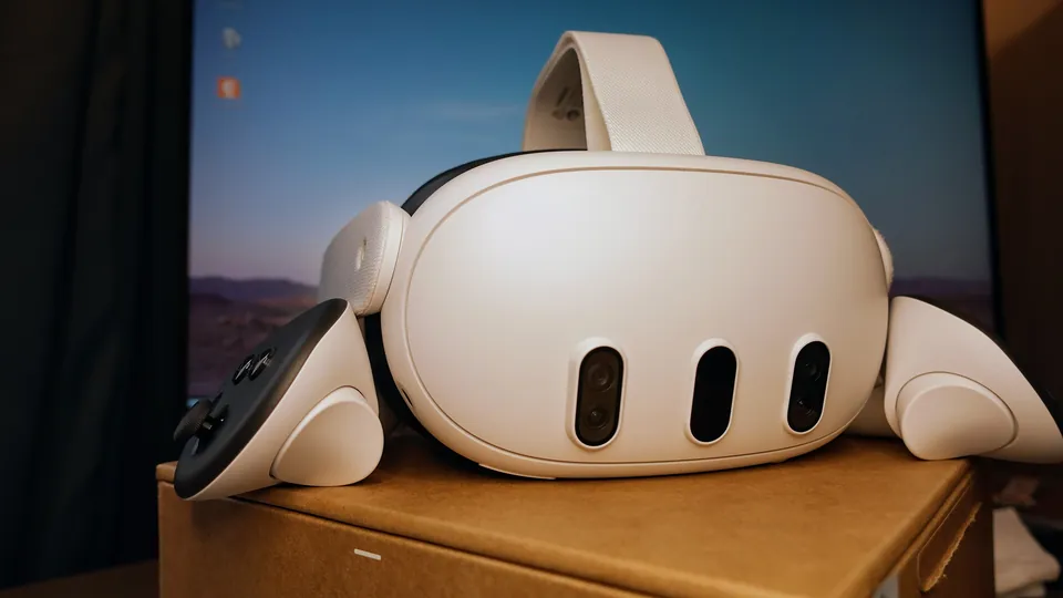 A white Meta Quest 3 headset with one controller, resting on a cardboard box in front of a monitor