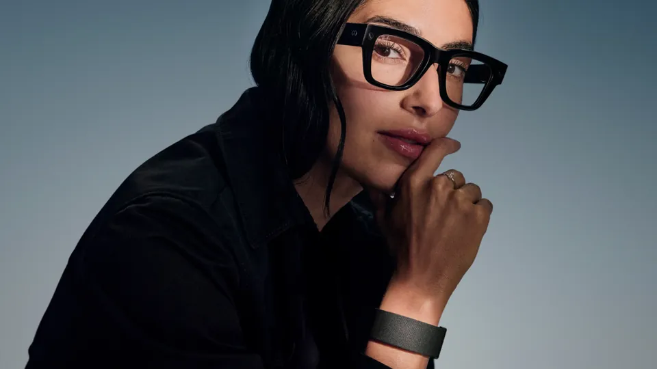 A person with dark hair in black-framed Meta Ray-Ban Display glasses and a black jacket rests their chin on one hand, with a dark Meta Neural Band on the wrist, against a blue-gray background