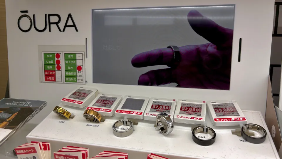 Oura rings in gold, rose gold, brushed silver, silver, black and stealth finishes on a store display under an Oura sign, with electronic price tags