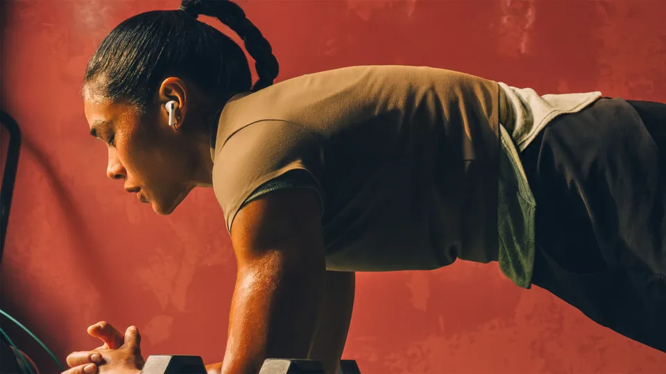 A woman with a braided ponytail holds a plank on a pair of dumbbells against a red wall, wearing an AirPods Pro 3 earbud in her ear