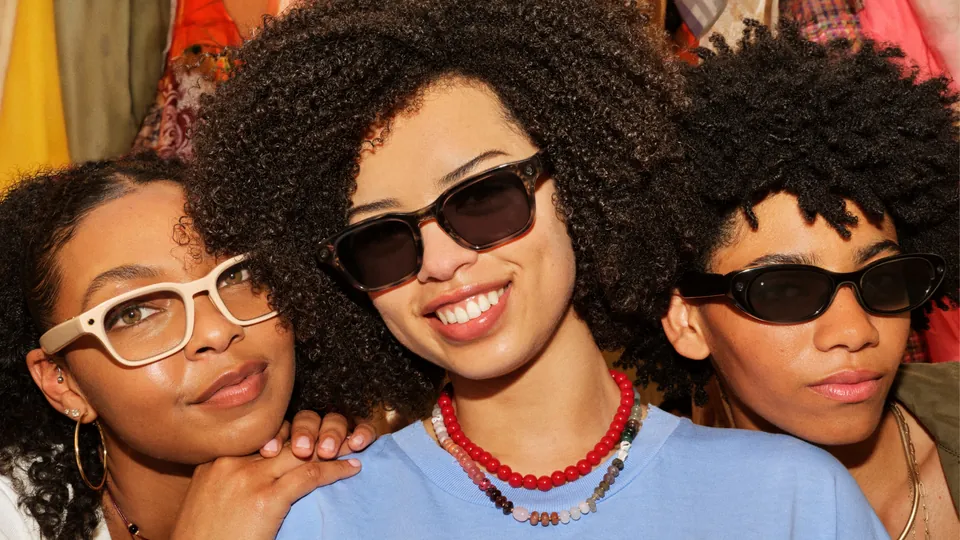 Three smiling friends with curly hair pose close together in a colorful room, wearing white-framed glasses, dark rectangular sunglasses and slim black oval sunglasses