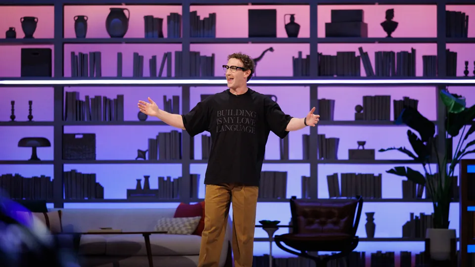 A man in a black T-shirt and tan trousers stands with his arms open on a stage in front of a lit wall of bookshelves