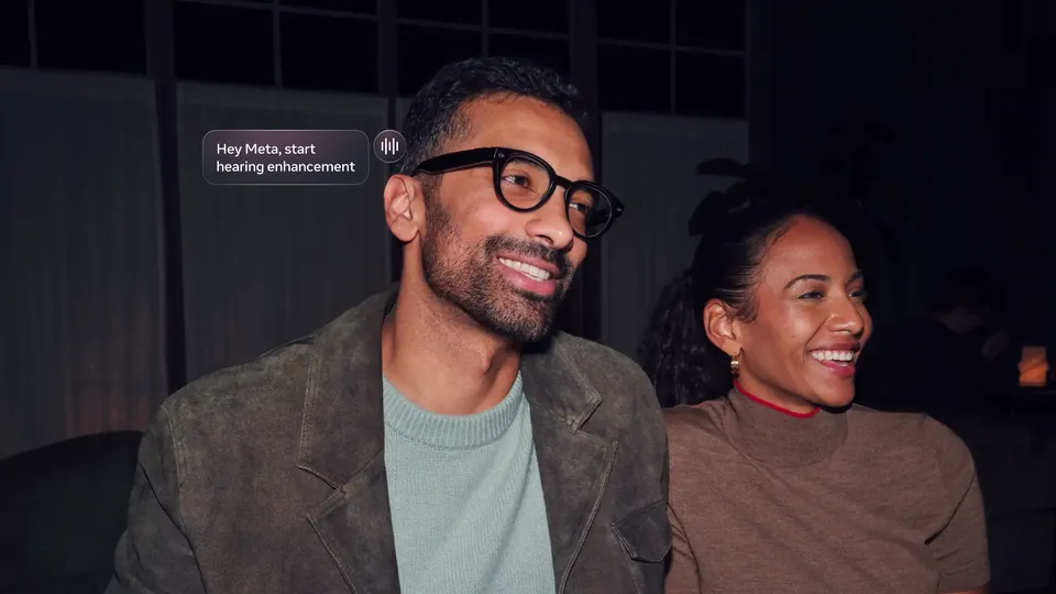 A smiling man in black round glasses and a gray-green sweater sits beside a smiling woman in a dim venue, with a message bubble reading Hey Meta, start hearing enhancement