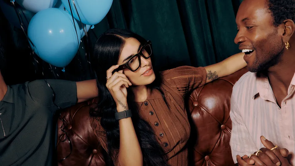A woman in thick black-framed glasses touches the frame and smiles toward a laughing man on a leather couch at a party with blue balloons, a Neural Band on her wrist