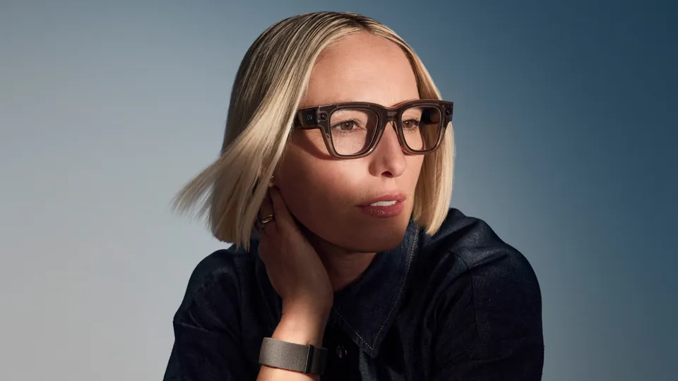 A woman with a blond bob wearing brown-framed Meta Ray-Ban Display glasses looks to the side with a hand at her neck, a gray Meta Neural Band on her wrist