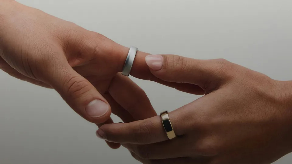 Two hands reaching toward each other against a pale gray background, one wearing a matte silver ring on the index finger and the other a polished gold-toned ring