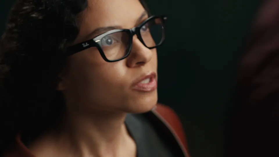 A woman with dark curly hair in black-framed glasses looks off to the side with a serious expression in a dim, dark-green space