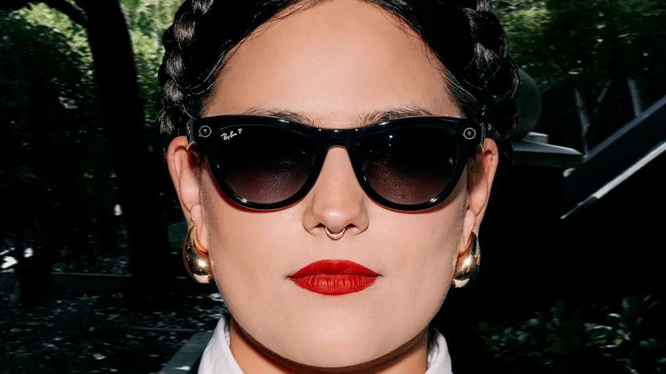 A woman with braided dark hair, gold earrings and red lipstick faces the camera in black cat-eye sunglasses with gradient lenses, a small camera lens at the corner of the frame