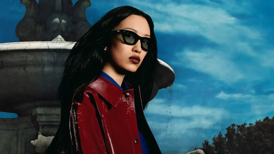 A woman with long dark hair in a glossy red jacket wears black rectangular glasses with green lenses in front of a stone fountain under a blue sky
