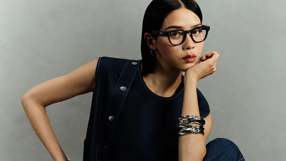 A woman with dark hair pulled back, in a dark denim vest and silver bracelets, rests her chin on her hand in round black-framed glasses against a gray wall