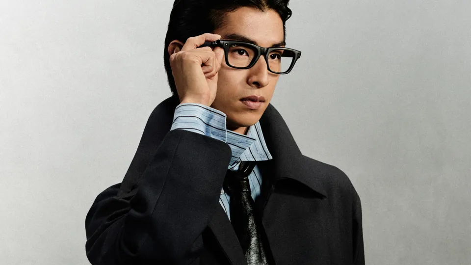A man in a dark coat, light blue shirt and tie adjusts the front of his black rectangular glasses with one hand against a gray wall