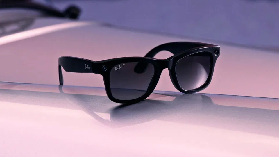 Black Ray-Ban Meta Wayfarer glasses with gradient gray lenses resting on a glossy pink and lilac surface, with a soft reflection beneath