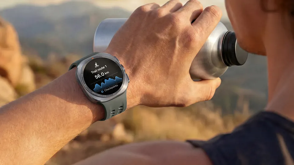 A runner drinks from a metal water bottle while wearing a Galaxy Watch Ultra2 with a green band, its screen showing Trail route 1 at 56.0 km with an elevation graph, against a blurred hillside