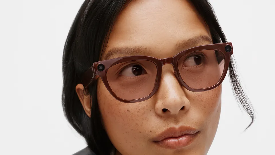 Close-up of a woman glancing to the side in glasses with a glossy brown frame and rose-tinted lenses, a small camera at each outer corner of the frame, against a white background
