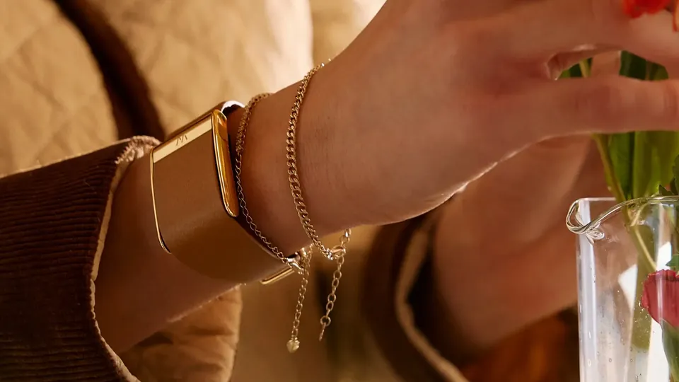 A wrist wearing a gold-toned WHOOP band on a LeatherLuxe strap beside two thin gold chain bracelets, the hand reaching toward a vase of flowers