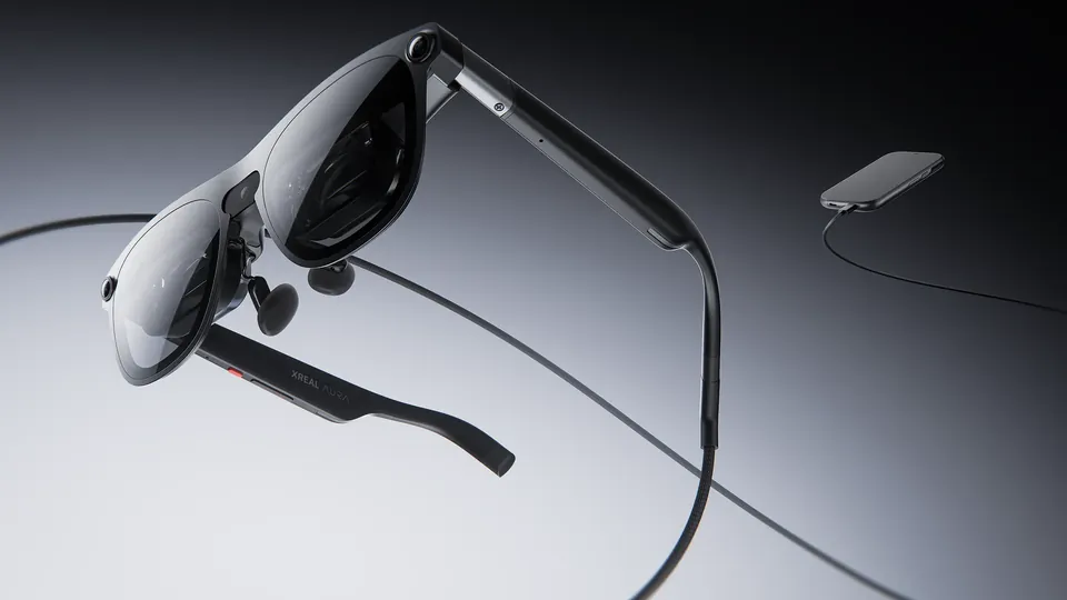 XREAL Aura glasses with dark lenses and a slim black temple floating over a gray studio backdrop, a braided cable looping away to a small compute puck at the upper right