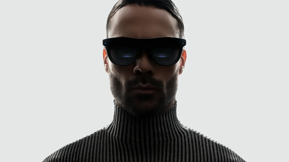A bearded man in a dark ribbed turtleneck faces the camera wearing black XREAL Aura glasses, against a light gray background
