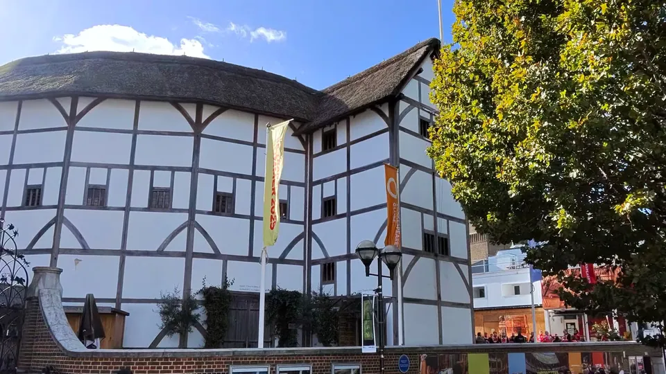 A white timber-framed building with a thatched roof, hanging flags and a tree beside it, under a blue sky