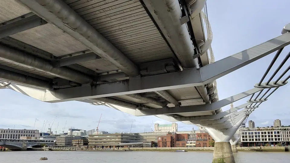 The underside of London's Millennium Bridge seen from the riverbank, with its steel deck and supports above the Thames and office buildings across the water