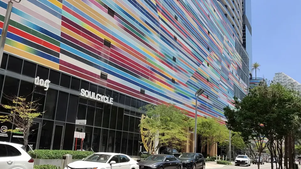 A tall building with a colorful striped facade above storefronts and parked cars on a sunny street in Miami's Brickell neighborhood