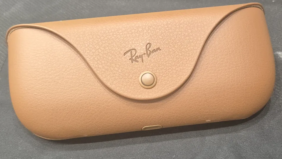 Closed tan Ray-Ban charging case for Ray-Ban Meta glasses lying on a dark fabric surface