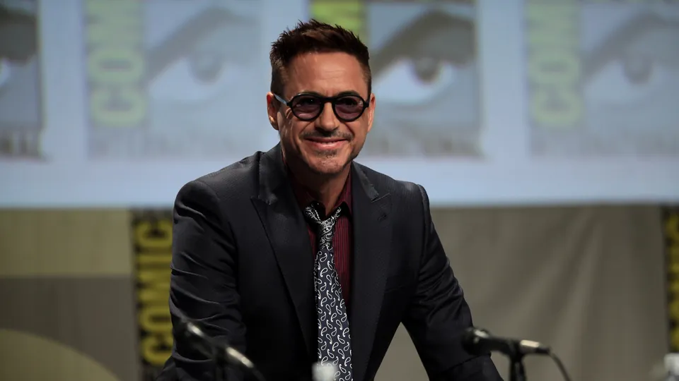 Robert Downey Jr., wearing round dark glasses and a suit, smiling on stage at a Comic-Con panel