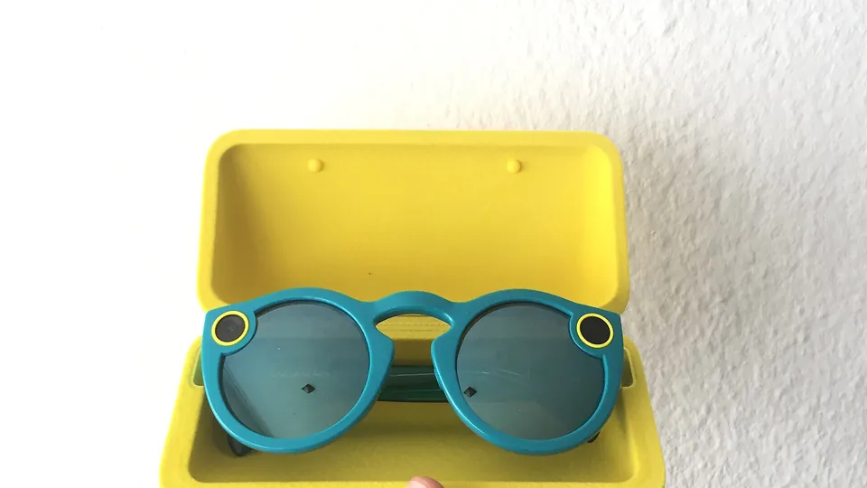 Teal first-generation Snap Spectacles sunglasses with a yellow-ringed circle at each front corner, sitting in their open yellow charging case