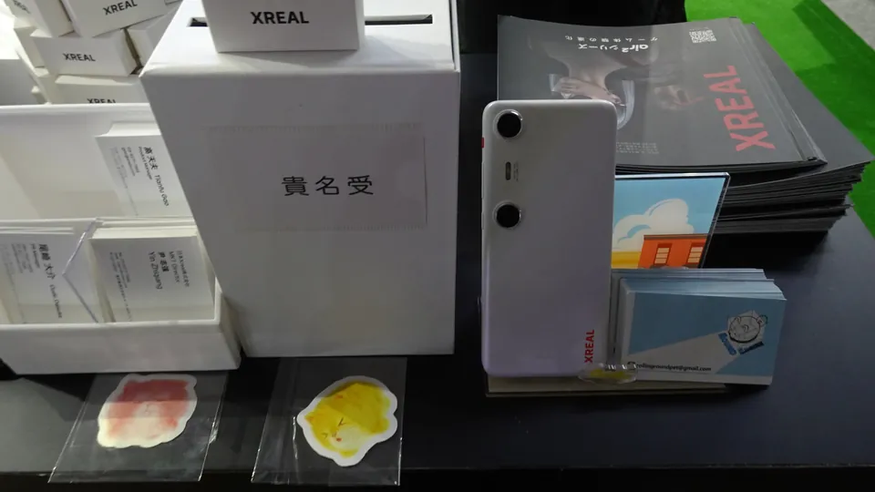 A white XREAL Beam Pro, a phone-sized device with two round camera lenses on the back and the XREAL name along its edge, standing on a table next to printed XREAL brochures and business cards