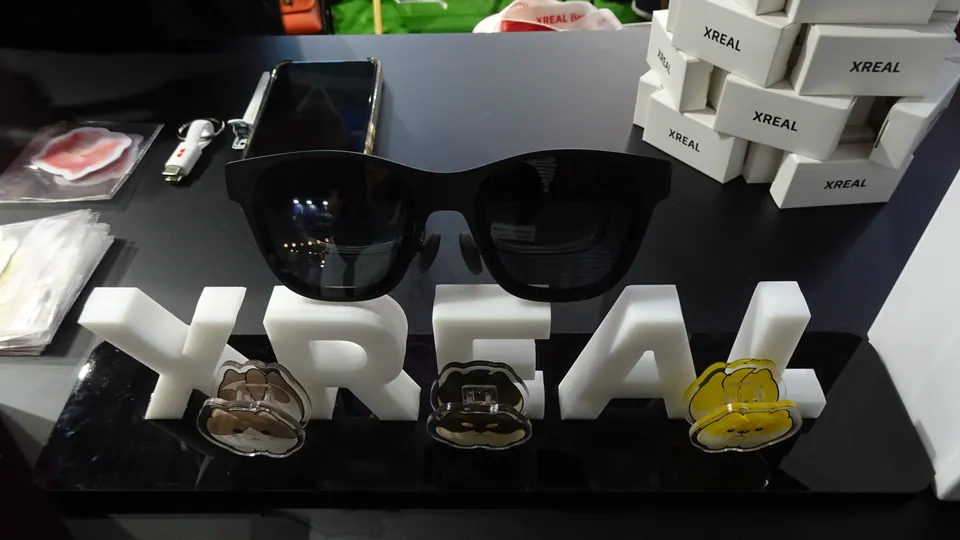 A pair of black XREAL glasses displayed on large white XREAL letters at a trade show booth, with boxes stacked behind